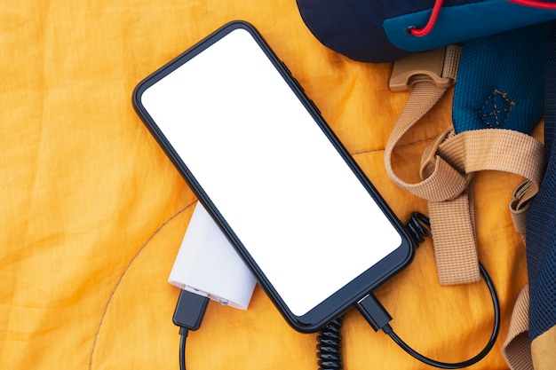 Smartfon ładuje się za pomocą przenośnej ładowarki. Power bank z makietą białego ekranu telefonu komórkowego na śpiworze z plecakiem.