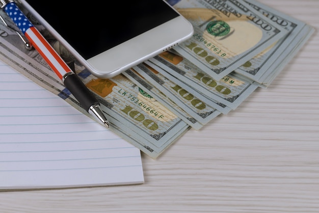 Smartphone, notatnik i pióro na sto dolarowych rachunkach.