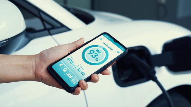 Smartphone wyświetla stan baterii w aplikacji mobilnej smart EV podczas ładowania samochodu elektrycznego elektrycznością przez domową stację ładowania w garażu Przyszły innowacyjny samochód elektryczny i zrównoważony rozwój energetyczny Przeczytaj