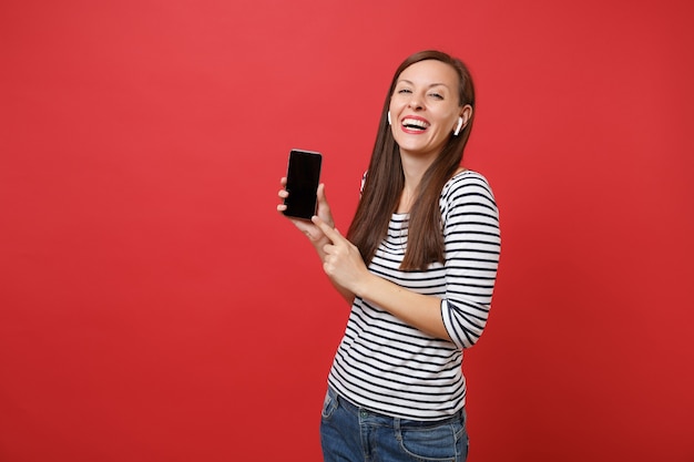 Śmiejąc się młoda kobieta z bezprzewodowymi słuchawkami trzymać telefon komórkowy z pustym czarnym pustym ekranie słuchania muzyki na białym tle na czerwonym tle. Ludzie szczere emocje, koncepcja stylu życia. Makieta miejsca na kopię.