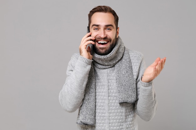 Śmiejąc się młody człowiek w szary sweter, szalik na białym tle na szarym tle portret studio. Zdrowy styl życia moda, emocje ludzi, koncepcja zimnej pory roku. Makieta miejsca na kopię. Rozmawia przez telefon komórkowy.