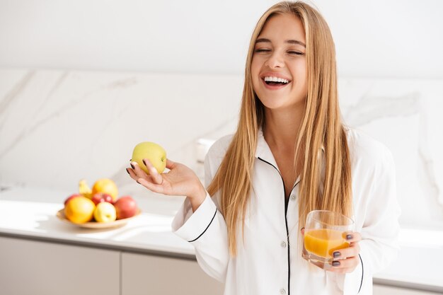 Śmiejąca Się Atrakcyjna Kobieta W Piżamie Trzymająca Jabłko I Pijąca Sok, Stojąc W Jasnej Kuchni
