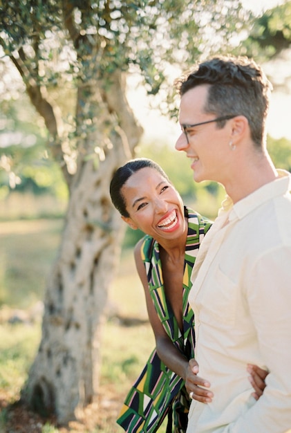 Śmiejąca się kobieta patrząca na mężczyznę przytulającego mu talię w pobliżu zielonego drzewa w ogrodzie