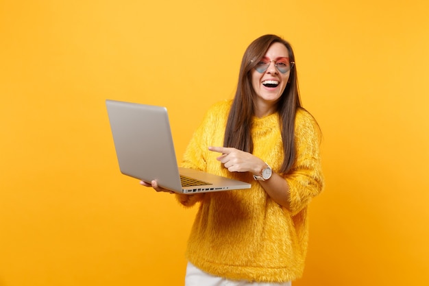 Śmiejąca Się Kobieta W Futrze Sweter, Serce Okulary Pracy, Wskazując Palcem Wskazującym Na Komputerze Typu Laptop Na Białym Tle Na Jasnym żółtym Tle. Ludzie Szczere Emocje, Koncepcja Stylu życia. Powierzchnia Reklamowa.