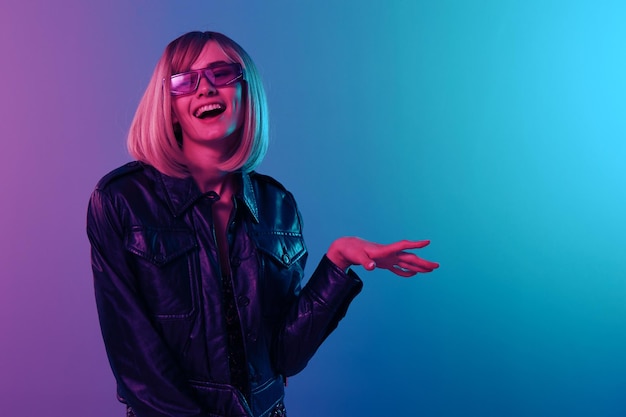 Śmiejąca się seksowna piękna blondynka w skórzanej kurtce błyszcząca sukienka modne okulary przeciwsłoneczne patrzą na bok pozują izolowane w niebiesko-różowym kolorze światło studia Neon party Cyberpunk koncepcja kopiowania przestrzeni