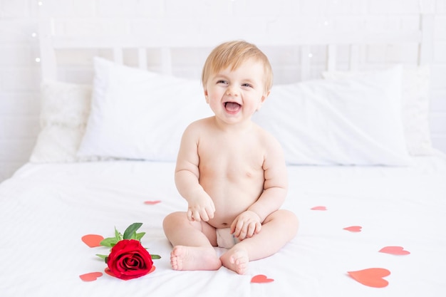 Śmiejący się blond chłopiec siedzi na łóżku w pieluchach i z czerwoną różą w domu i uśmiecha się między sercami koncepcja Dnia Walentynek lub Dnia Matki