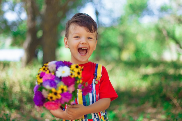 Śmiejący się chłopiec z bukietem kwiatów dziecko z bukietem dzikich kwiatów dla mamy prezent niespodzianka własnymi rękami emocjonalne dziecko z kwiatami w jasnych ubraniach w naturze