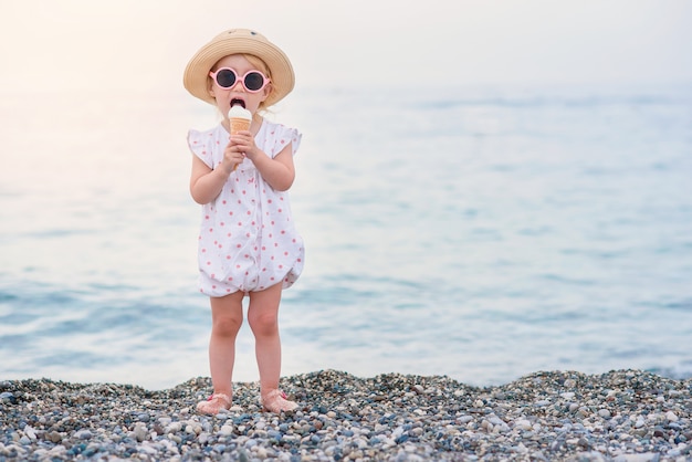 Śmieszna Berbeć Dziewczyna Jest Ubranym Różowe Letnie Kombinezony, Kapelusz I Różowe Okulary Przeciwsłoneczne Je Waniliowe Białe Lody Podczas Wakacji Na Plaży.