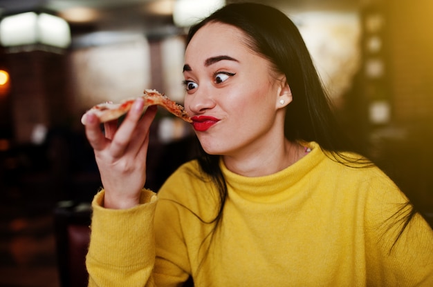 Śmieszna brunetki dziewczyna w żółtej puloweru łasowania pizzy przy restauracją.