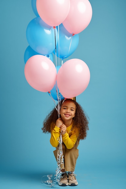 Śmieszna Dziewczynka W Czapce Trzyma Kilka Kolorowych Balonów, Niebieskie Tło. ładne Dziecko Dostało Niespodziankę, Imprezę Lub Przyjęcie Urodzinowe