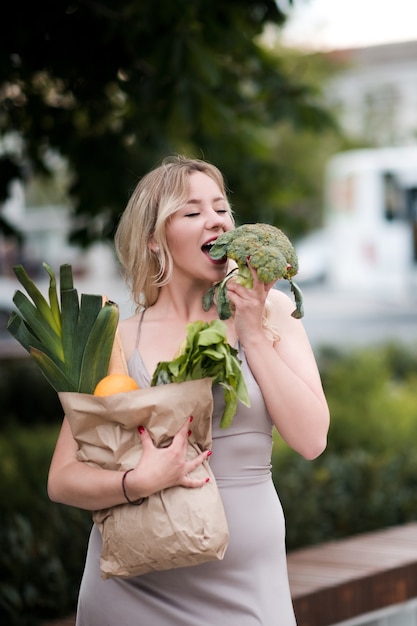Śmieszna kobieta trzymająca papierową torbę z jedzeniem gryzie świeże zielone brokuły