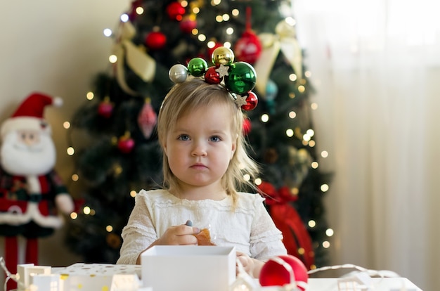 Śmieszna mała dziewczynka w białej sukni otwierającej świąteczne pudełko w domu w pobliżu choinki