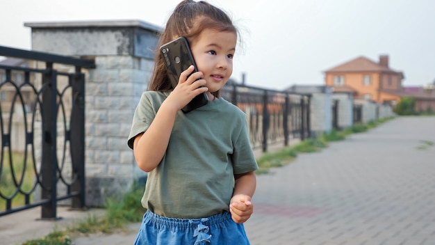 Śmieszna mała koreańska dziewczyna z długim kucykiem mówi na nowoczesny telefon komórkowy stojący na nabrzeżu pustego miasta o zachodzie słońca blisko widok