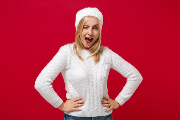 Zdjęcie Śmieszna młoda kobieta w białym swetrze kapeluszu odizolowywającym na czerwonym ściennym tle portretu pracownianym. zdrowy styl życia mody, koncepcja zimnej pory roku. makieta miejsca na kopię. stań z rękami opuszczonymi wokół talii, mrugając.