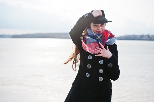 Zdjęcie Śmieszna przypadkowa młoda dziewczyna przy czarnym żakietem, szalikiem i kapeluszem przeciw zamarzniętej rzece przy pogodną zimy pogodą.
