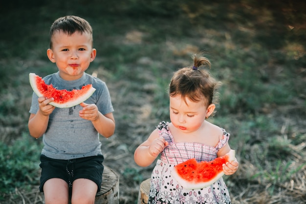 Śmieszne dzieci jedzą arbuza. Brat i siostra na świeżym powietrzu, siedząc na pniakach