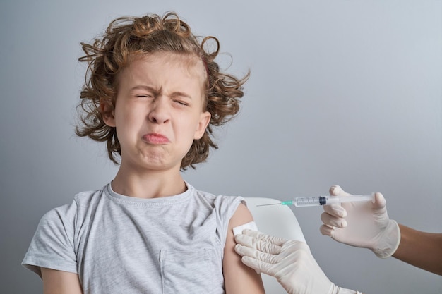 Śmieszne, przestraszone dziecko w szarej koszulce z mocno zamkniętymi oczami i ustami, przygotowujące się do szczepienia