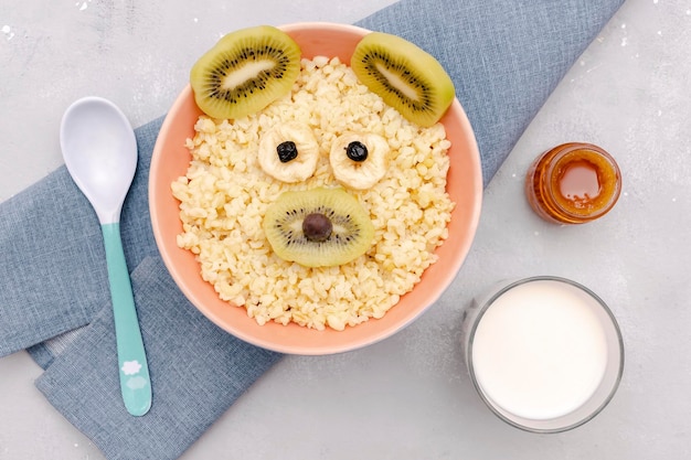 Śmieszne słodkie dzieci dla dzieci zdrowe śniadanie obiadowe płatki owsiane w misce wyglądają jak twarz myszy niedźwiedziej ozdobione kiwi bananem suszone owoce jagodowemleczny deser jedzenie sztuki na szarym stole