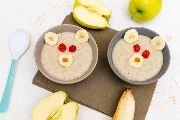 Śmieszne słodkie dzieci dla dzieci zdrowe śniadanie obiadowe płatki owsiane w misce wyglądają jak twarz niedźwiedzia ozdobiona jabłkiem bananem suszone owoce jagodowe deser sztuka żywności na białym drewnianym stole