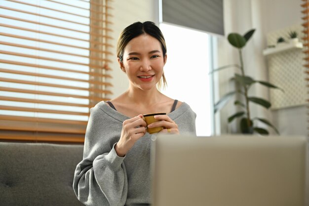 Smilie tysiącletnia kobieta pijąca herbatę ziołową cieszy się wolnym weekendem w domu, przeglądając internet na laptopie