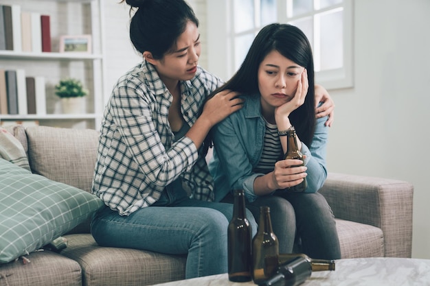 smutna kobieta pije alkohol w domu, siedząc na kanapie na kanapie w jasnym, nowoczesnym salonie z dużą ilością pustych butelek na stole. młoda dziewczyna uwalnia ciśnienie piwem. azjatycki dama pocieszając swojego przyjaciela.