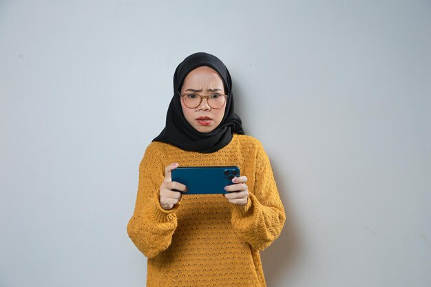 smutna młoda azjatycka muzułmanka ubrana w pomarańczowy sweter i okulary grająca w grę na telefonie komórkowym