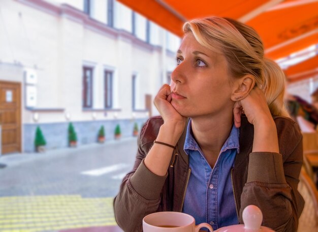 Smutna młoda blondynka siedzi w kawiarni z filiżanką herbaty i w zamyśleniu patrzy w bok Orientacja pozioma selektywna ostrość przestrzeń kopiowania