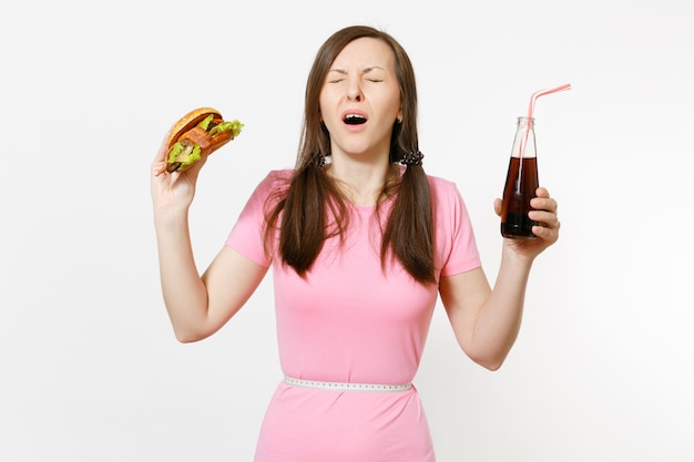 Smutna młoda kobieta z ogonami, miarką w pasie, trzymając burgera, colę w szklanej butelce na białym tle. Prawidłowe odżywianie lub amerykańskie klasyczne fast foody. Powierzchnia reklamowa z miejscem na kopię.