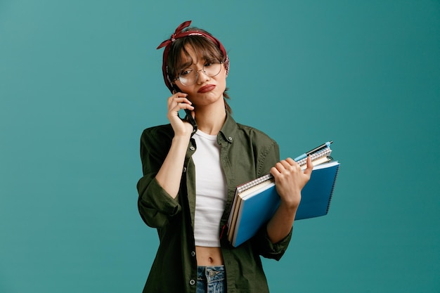 Smutna młoda studentka w okularach bandana, trzymająca duże notatniki z długopisem, patrząca w bok podczas rozmowy przez telefon na białym tle na niebieskim tle