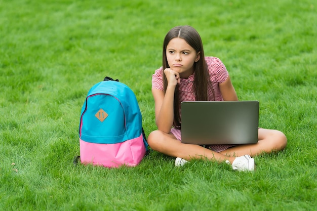 Smutna nastolatka korzystająca z notebooka do edukacji online i nauki, siedząca w parku na zielonej trawie z plecakiem, wraca do szkoły.