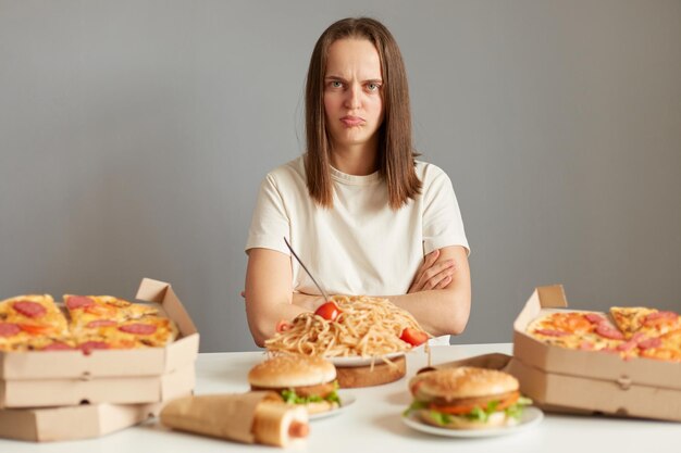 Smutna zdenerwowana nieszczęśliwa kobieta z brązowymi włosami ubrana w białą koszulkę siedząca przy stole wśród niezdrowej żywności odizolowanej na szarym tle wygląda na urażona siedząc z założonymi rękami