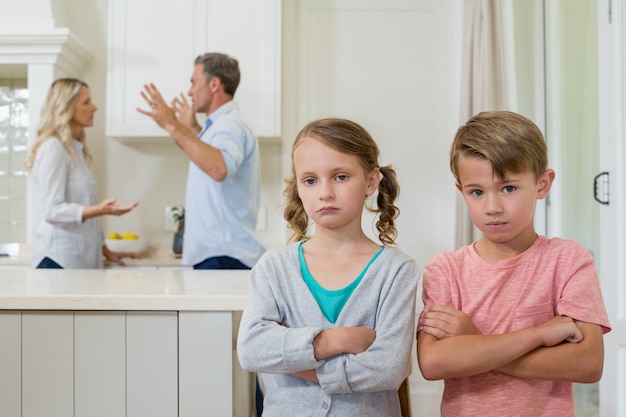 Smutne rodzeństwo stojące ze skrzyżowanymi rękami, podczas gdy rodzice się kłócą