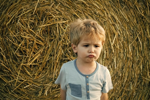 Smutny chłopiec przy beli siana, lato. Małe dziecko bawi się na farmie lub polu ranczo, wakacje. Agroturystyka, ekoturystyka, wypoczynek, podróże. Lato, żniwa. Wzrost młodości w dzieciństwie