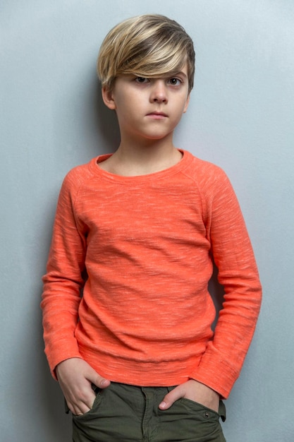 Smutny chłopiec w wieku 910 lat stoi przy szarej ścianie Facet w pomarańczowym swetrze trzyma ręce w kieszeniach Samotność i depresja Pionowo