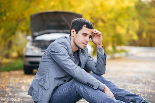 Smutny mężczyzna siedzi na drodze przed zepsutym samochodem z otwartą maską