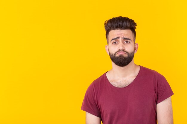 Smutny, młody przystojny mężczyzna hipster z brodą w bordowej koszulce pozowanie na żółtej powierzchni. Pojęcie frustracji. Przestrzeń reklamowa