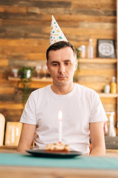 Smutny, samotny młody człowiek w świątecznym kapeluszu świętujący urodziny sam, siedzący na urodzinowym torcie i patrzący na niego smutnymi oczami. Koncepcja świętowania sam.