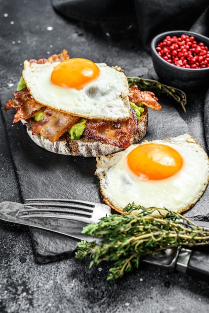 Śniadanie angielskie, tost z bekonem, awokado i jajkiem na desce do krojenia. Zdrowe jedzenie. Czarne tło. Widok z góry.