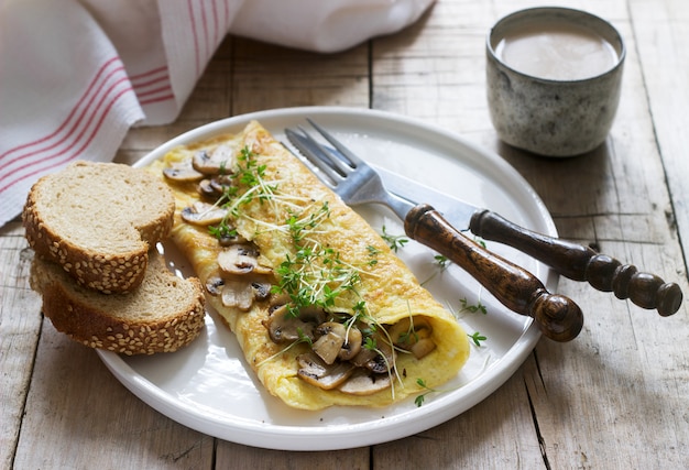 Śniadanie wegetariańskie, omlet z pieczarkami i rzeżuchą, podawane z chlebem żytnim i kawą z mlekiem. Styl rustykalny.