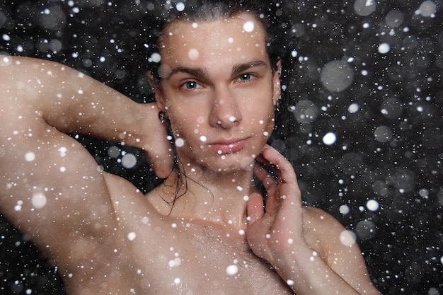 Śnieg, zima, boże narodzenie, ludzie, pielęgnacja skóry i koncepcja urody - mokry młody człowiek z długimi czarnymi włosami na czarnym tle śniegu. Portret mężczyzny z ogoloną klatką piersiową. Pielęgnacja męskiej skóry.