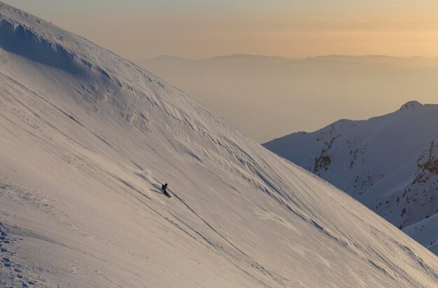 Snowboardzista schodzi szerokim zboczem na tle pasm górskich i zachodu słońca