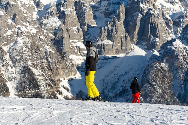 Snowboardzista w czarnej kurtce i żółtych spodniach stoi na stoku narciarskim na tle góry.