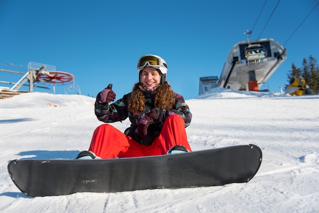 Snowboardzistka dziewczyna siedzi na śniegu w pobliżu wyciągu narciarskiego na tle błękitnego nieba i pokazuje kciuki w górę gesty dobrej klasy