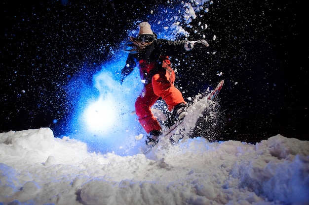 Snowboardzistka ubrana w czerwoną odzież sportową skacząca na zboczu góry w nocy pod niebieskim światłem
