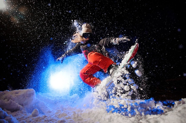 Snowboardzistka ubrana w pomarańczową odzież sportową, skacząca po zboczu góry w nocy pod niebieskim światłem