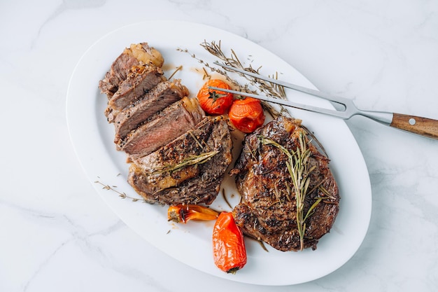 Soczyste, dobrze usmażone steki ribeye na białym talerzu na białej marmurowej powierzchni z malinami i smażonymi pomidorkami koktajlowymi oraz steki z papryki są pokrojone i gotowe do podania