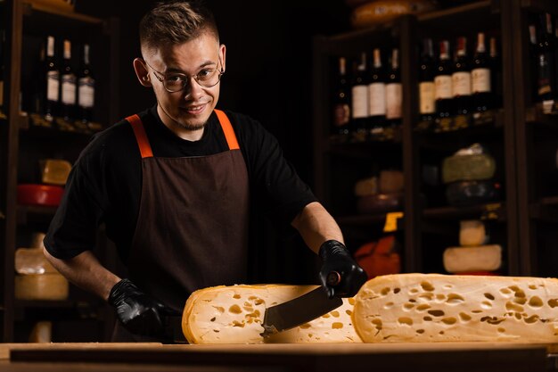 Sommelier serowy w sklepie spożywczym pracownik tnie ograniczony maasdam naturalny w wieku i sprawdza jakość Przystojny brodaty mężczyzna pracujący w sklepie spożywczym