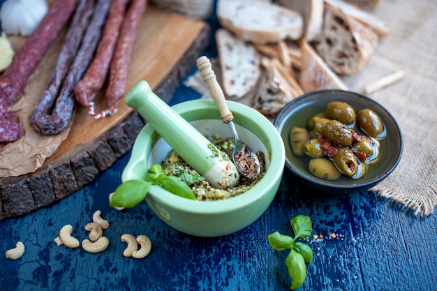 sos pesto w zielonej filiżance i różne salami na drewnianej desce, oliwki i niebieska powierzchnia