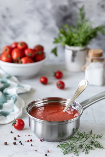 Sos świeżych czerwonych pomidorów w szklanym słoju na czarnym talerzu. Gałązka świeżych pomidorów cherry, czosnku, ostrej papryki, koperku i natki pietruszki na białym stole. Puszka domowego keczupu. Zbliżenie