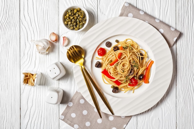 Spaghetti alla caponata z makaronu pełnoziarnistego z pomidorami, oliwkami, słodką czerwoną papryką i kaparami na białym talerzu na teksturowanym białym drewnianym stole, płasko leżąc